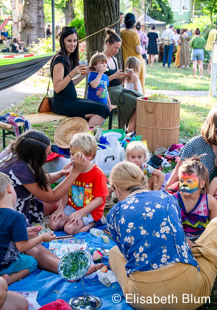 Several kids at an outdoor children station geting their faces painted. Some mothers are watching.