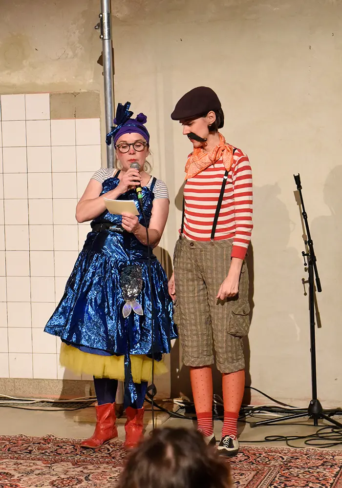 The two moderators of Salon Sardine on the stage. One wears a glittering dress, the second is dressed in a striped shirt and trousers with a checked pattern, has a moustache and wears a cap